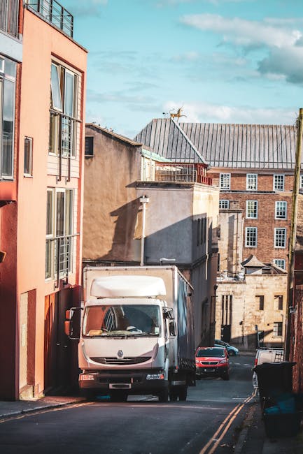 An urban scene featuring a street in Mill Hill with a row of red-brick residential buildings, some with decorative turrets topped with dome-shaped roofs and small wind vanes. The buildings have multip