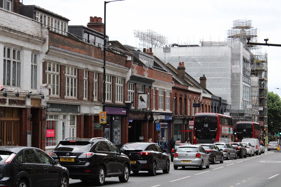 A row of two-story townhouses and commercial buildings on Mill Hill Broadway, with brick facades and large sash windows, situated along a busy urban street. Several black, grey, and white cars are parked along the roadside, with some moving traffic visible in the background. Two red double-decker buses numbered 343 and 42 are seen traveling along the street, indicating active public transport. Construction scaffolding and white protective sheeting partially cover a building at the far end of the row, suggesting ongoing renovation work. The pavement is populated with pedestrians, and a street sign indicates a speed limit of 20 mph. Street lighting, traffic signals, and various shopfronts contribute to the lively commercial atmosphere. This scene exemplifies an active street environment encountered during home relocation or furniture transport activities managed by [COMPANY_NAME], illustrating typical urban move logistics involving parking, loading, and public transport integration near residential and retail premises.