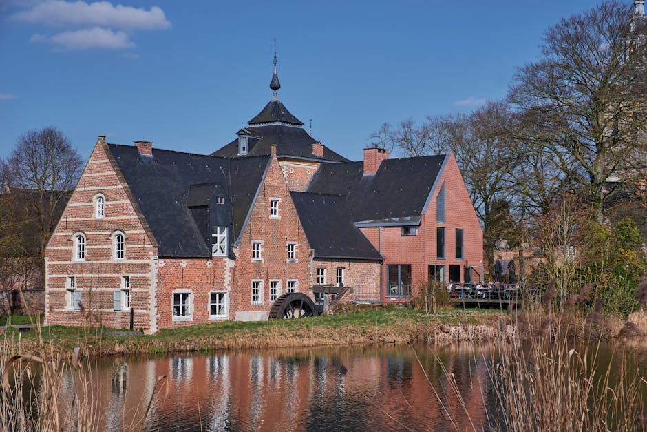A view of a narrow river flowing gently through a rural area, with lush green trees and foliage on both sides. On the right bank, there is an old wooden watermill structure with weathered dark brown timber walls and a sloped roof, positioned close to the water's edge. The watermill includes a large wheel, partially visible, connected to the building, indicating its historical use for milling or water management. A small stone bridge spans the river further in the background, blending into the natural setting. The sky above is clear and blue, and the scene is well-lit, highlighting the textures of the wooden watermill, the greenery, and the rippling surface of the river. This peaceful environment reflects a traditional rural landscape, suitable for illustrating themes related to property relocation, moving logistics, or rural house removals, with [COMPANY_NAME] providing professional services in house removals and furniture transport in the Mill Hill NW7 area.