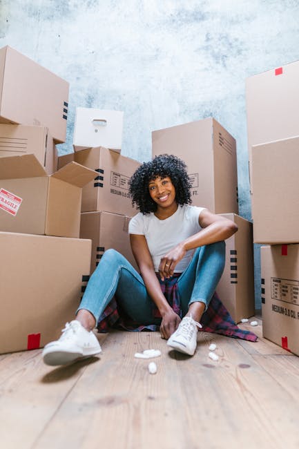 A woman with curly black hair and a bright smile is seated on a wooden floor surrounded by stacked cardboard moving boxes of various sizes, some with visible labels and packing tape. She is dressed in a white t-shirt, blue jeans, and white sneakers. A plaid blanket or cloth is draped around her waist. The setting appears to be inside a property with a light blue, slightly textured wall in the background, indicating a home environment ready for a house removal. The boxes are arranged closely together, and some are partially open or sealed with red tape, suggesting an active packing or unpacking process. Man and Van Mill Hill, specialists in removals, would likely handle the logistics of such home relocation, including the careful packing and transport of furniture and belongings during the moving process. The overall scene emphasizes preparation and organization in a domestic setting for furniture transport and moving services.