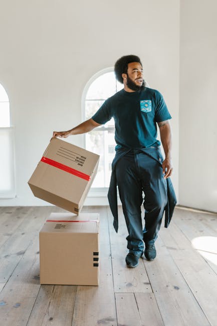 A man with a beard and dark curly hair is inside a bright, spacious room with light-colored walls and wooden flooring, preparing for a home relocation. He is wearing a navy blue t-shirt with a logo on the chest, dark trousers, and has a sweatshirt tied around his waist. He is holding a medium-sized cardboard box with red tape securing the top, about to place it on top of a larger box on the floor. Behind him, large arched windows allow natural light to illuminate the area. Nearby, additional cardboard boxes and packing materials are visible, indicating an ongoing packing and moving process. This scene reflects the logistics involved in furniture transport and packing for a residential move, and is associated with professional removals conducted by [COMPANY_NAME], as part of their comprehensive house removals services, highlighting the careful handling of packing and loading during the transition from Mill Hill East to The Hale.