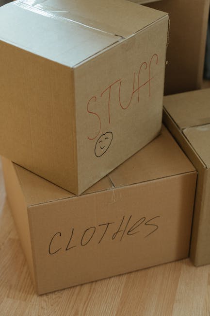 A woman with curly black hair and a bright smile is seated on a wooden floor surrounded by stacked cardboard moving boxes of various sizes, some with visible labels and packing tape. She is dressed in a white t-shirt, blue jeans, and white sneakers. A plaid blanket or cloth is draped around her waist. The setting appears to be inside a property with a light blue, slightly textured wall in the background, indicating a home environment ready for a house removal. The boxes are arranged closely together, and some are partially open or sealed with red tape, suggesting an active packing or unpacking process. Man and Van Mill Hill, specialists in removals, would likely handle the logistics of such home relocation, including the careful packing and transport of furniture and belongings during the moving process. The overall scene emphasizes preparation and organization in a domestic setting for furniture transport and moving services.