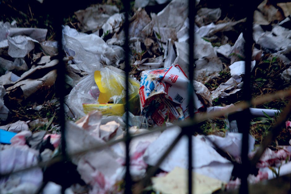 A black cylindrical public waste bin with a domed lid, positioned on a cobbled street surface in an urban setting. The bin's surface and surrounding area are cluttered with litter, including several g