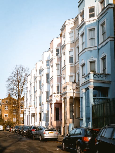 A vintage red and white Volkswagen van is parked on a residential street in Mill Hill, next to a row of colorful Victorian-style terraced houses with decorative facades and bay windows. The van is pos